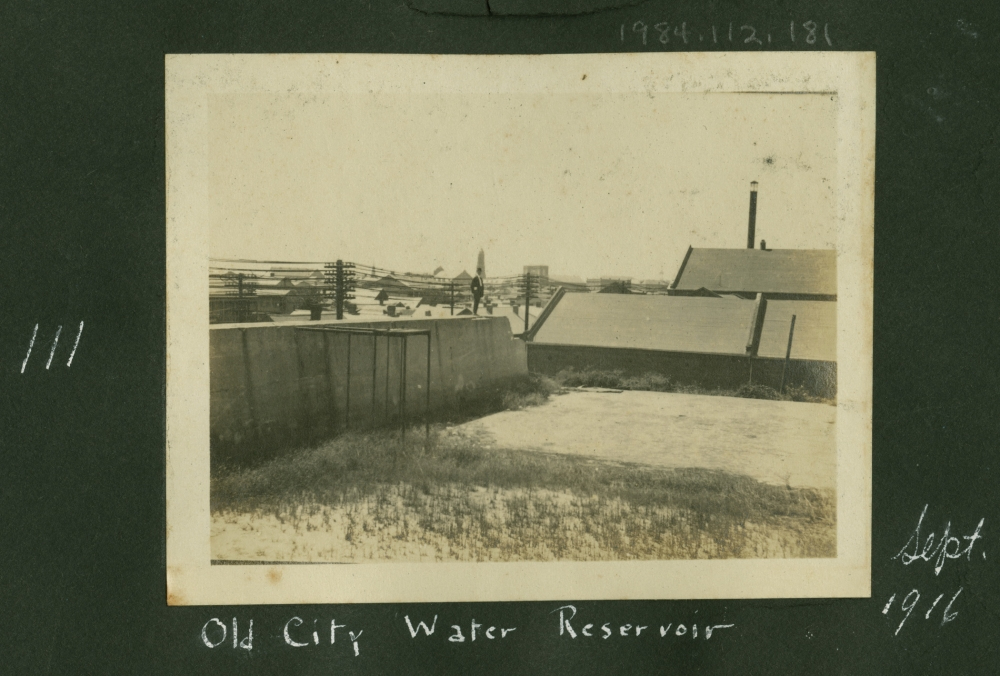A vintage photograph of an old city water reservoir dated September 1916. The image shows a large concrete structure surrounded by grass, with rooftops and a chimney visible in the background. Handwritten notes are visible along the edges.