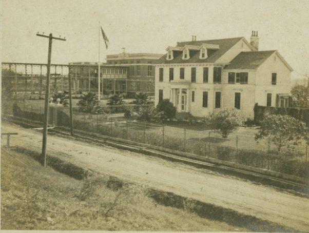A sepia-toned vintage photograph of a large residence with a porch, surrounded by a fence and garden. A flag flies on a pole nearby, and a train track runs parallel to a dirt road in the foreground.