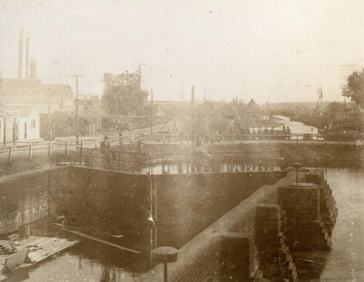 A sepia-toned historical photo showing an industrial canal lock. People and structures line the canal, with smokestacks and trees in the background. The waterway runs through the composition, surrounded by brickwork and fencing.