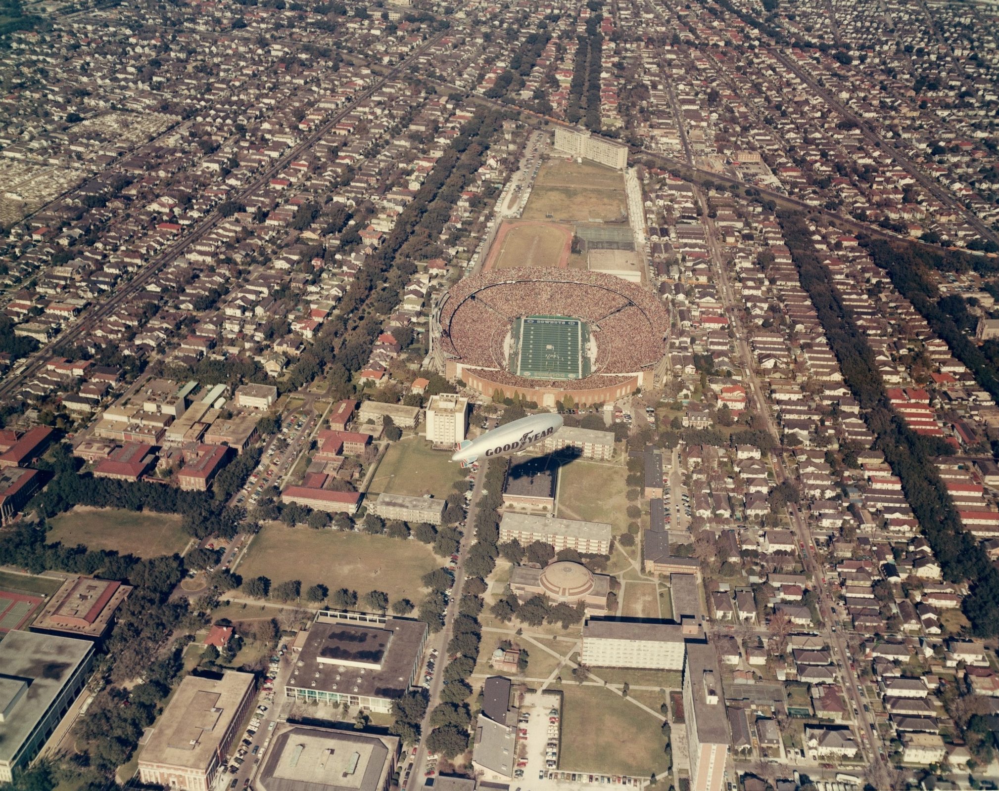 Aerial view of a densely built city featuring a crowded stadium with a visible sports field in the center. Surrounding the stadium are numerous buildings and streets laid out in a grid pattern.