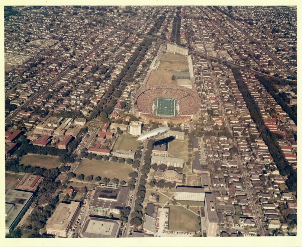 Aerial view of a large football stadium filled with spectators. The stadium features a green field and is surrounded by densely packed urban streets and buildings. A blimp hovers above, prominently displaying advertising.