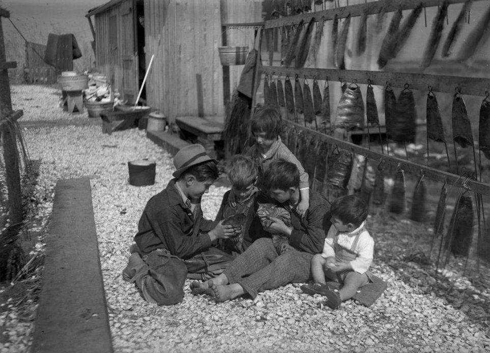 Five children sit on gravel outside, engrossed in looking at something held by one of them. Theyre dressed in rustic attire. Behind them are wooden structures, and fish are hanging on a wooden rack to dry. The setting appears rural and historical.