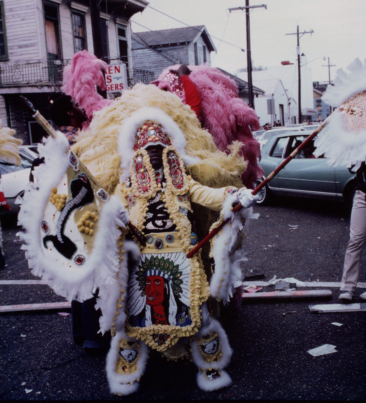 A Mardi Gras Indian dressed in a vibrant, elaborate costume with feathers and beadwork stands on a street. The outfit features a decorative central panel with an intricate design. Buildings and a slightly overcast sky form the background.