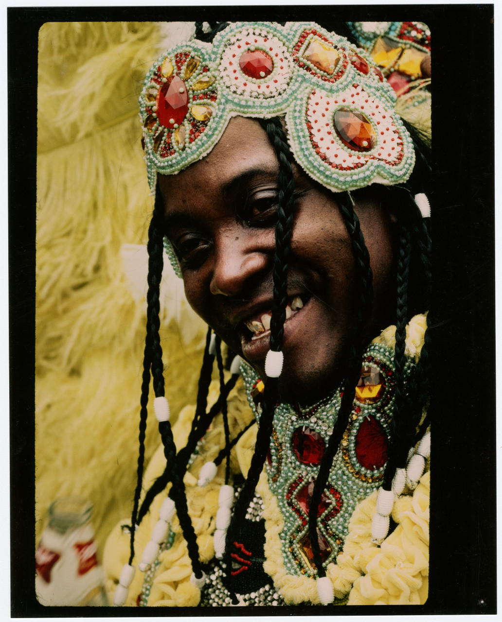 A person smiles broadly, dressed in a vibrant, intricately beaded costume with braided hair adorned with beads. The outfit includes colorful gemstones, feathers, and detailed patterns, creating a festive and lively appearance.