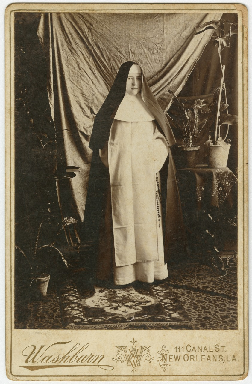 A sepia-toned vintage photograph of a nun in traditional habit. She stands in a room with a patterned carpet and draped curtain backdrop. Potted plants and a small table with a vase can be seen in the background.