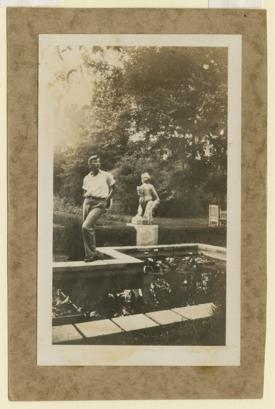 Vintage photograph of a man wearing a white shirt and slacks, standing near a statue and a backyard pond in the 1930s.