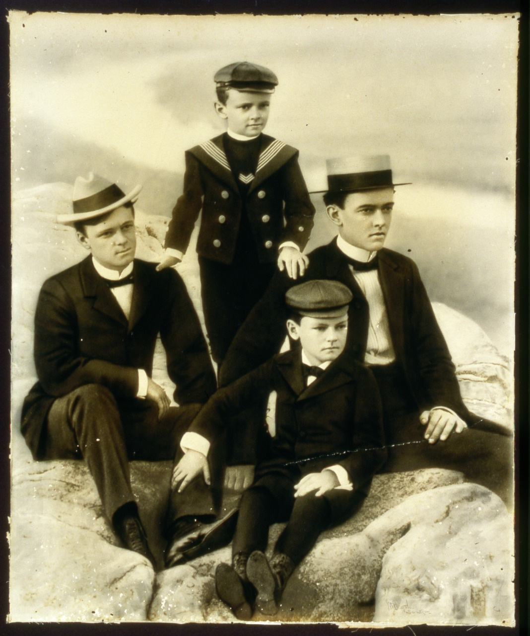 A vintage photograph of four boys seated on rocks, each wearing period attire including jackets, hats, and bow ties. The tallest boy is standing, while the others sit. The background is softly blurred, suggesting a natural setting.