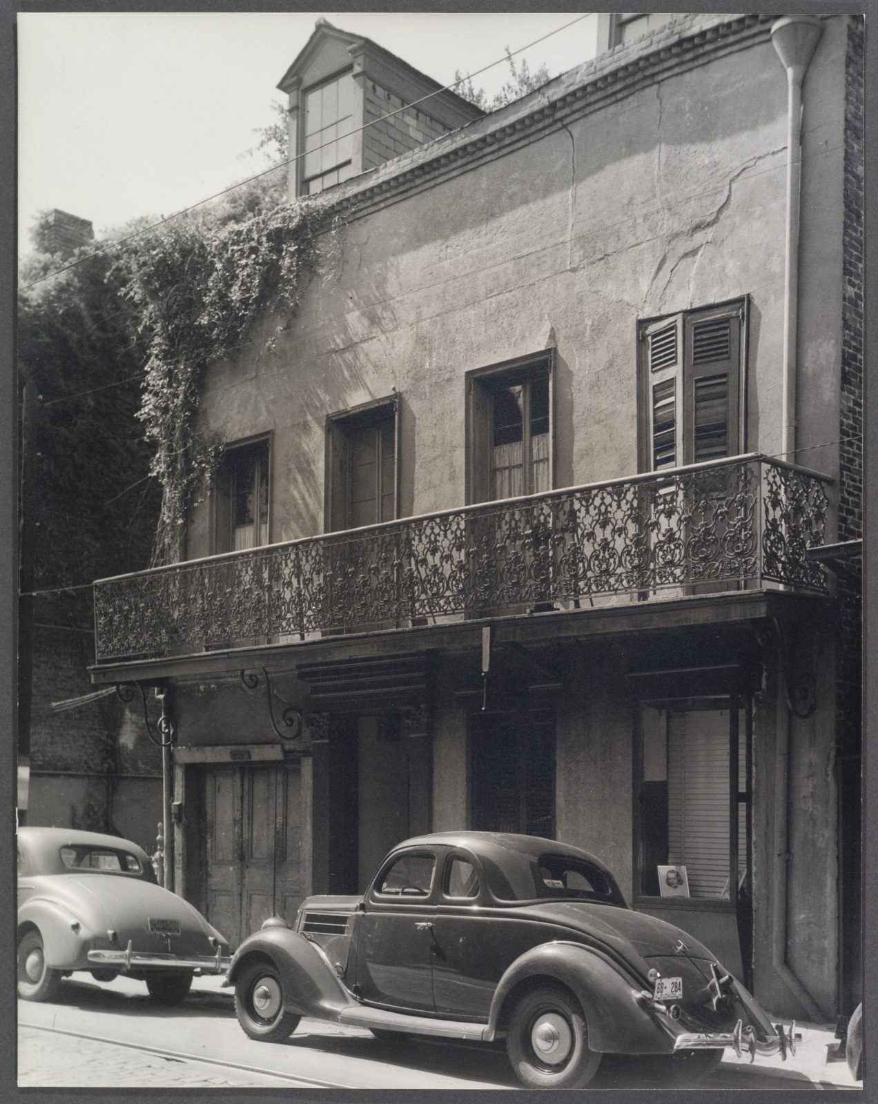 A vintage black-and-white photo shows two classic cars parked in front of an old building with shuttered windows and an ornate wrought-iron balcony. The upper floor has some ivy growing on it, adding charm to the scene.