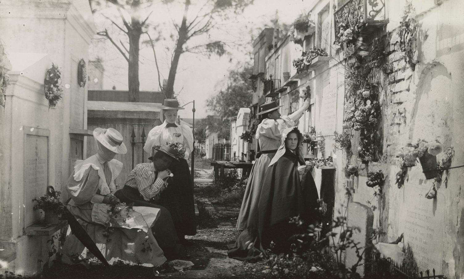 A black and white image of four women tending to above-ground graves in a cemetery. Some are cleaning the tombs, while others arrange flowers. The scene is set along a narrow path lined with trees in the background.