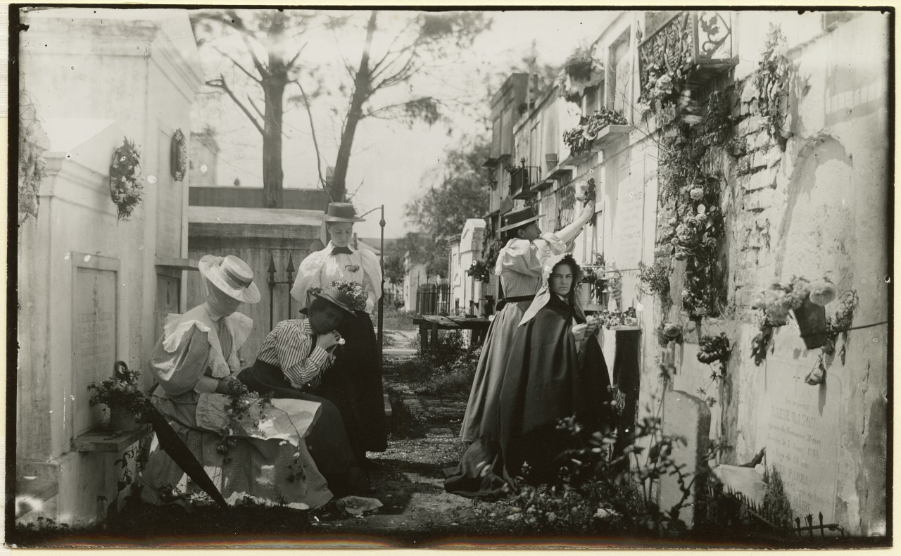 People gather, tending to graves in a cemetery adorned with flowers. A seated woman holds an umbrella, another adjusts flowers, while others stand nearby, dressed in early 20th-century attire. The atmosphere is somber and respectful.