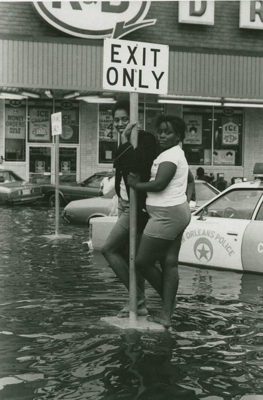 Two women pose on a pole labeled Exit Only in a flooded street. Behind them, a New Orleans Police car is partially submerged. The scene is in front of a storefront with visible signs for ice and prices.