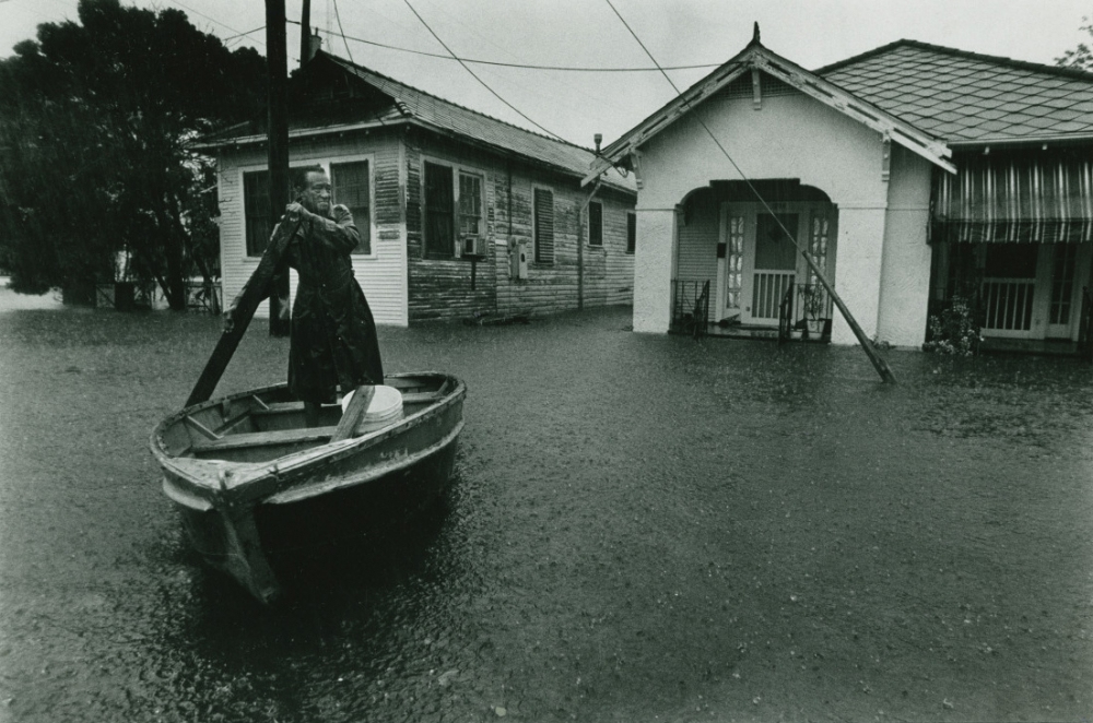 A person stands in a small boat navigating floodwaters in a residential area. The water is high, reaching up to the doors of wooden houses. The sky is overcast, and power lines are visible. The image is in black and white.