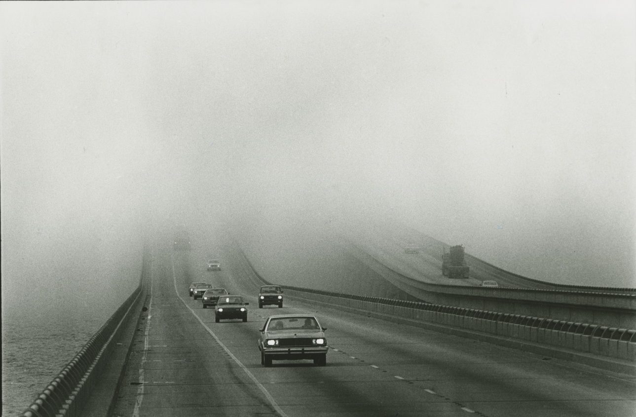 A black and white photo shows cars driving out of a cloudbank over the Lake Pontchartrain Causeway, taken in 1983 or 1984.