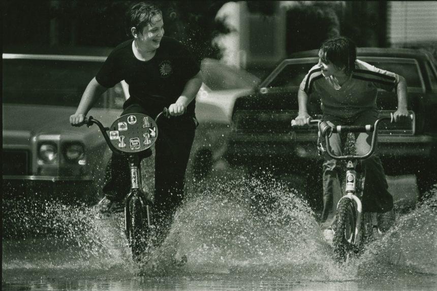 Two people joyfully ride bicycles through a large puddle on a street. They are splashing water and smiling at each other. Cars are parked along the road in the background.