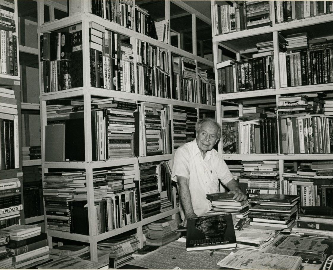 An elderly person stands among towering bookshelves filled with books, wearing a white shirt. The scene suggests a library or a personal collection. They look content, surrounded by a variety of books.