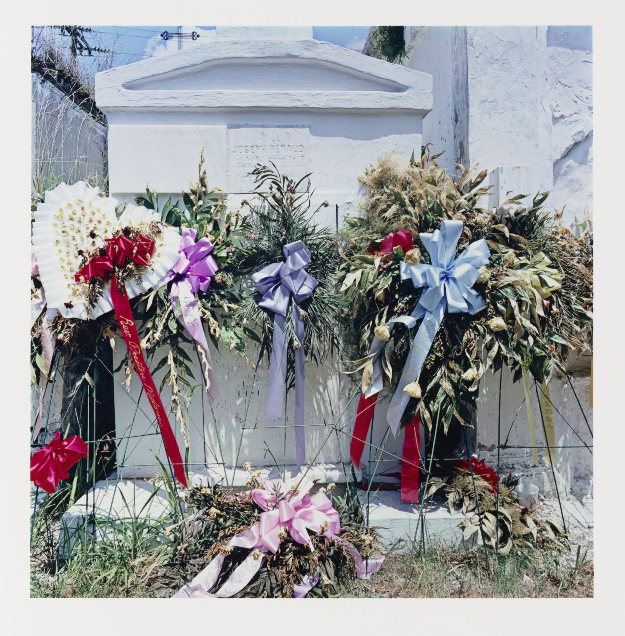 Floral wreaths with colorful ribbons adorn an old stone monument. The background shows weathered inscriptions and foliage, with a few tufts of grass in the foreground. The scene suggests a somber, commemorative atmosphere.
