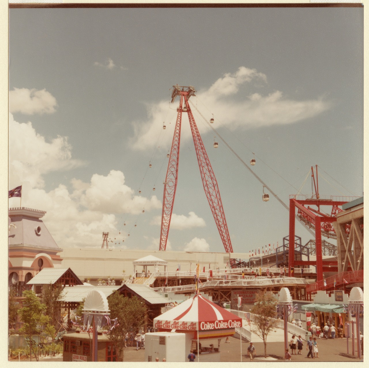 A vibrant amusement park scene under a clear sky featuring a tall red swing ride with gondolas, a striped Coke tent, various flags, and crowds of people enjoying the sunny day.