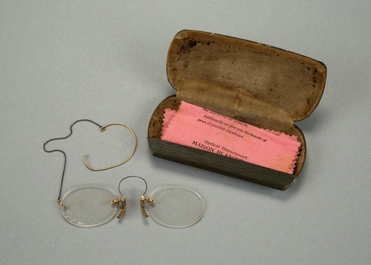 Vintage pince-nez glasses with oval lenses and a nose clip, accompanied by a chain and ear loop. Includes a pink cleaning cloth from Maison Bijou Optical Department, housed in a brown case.