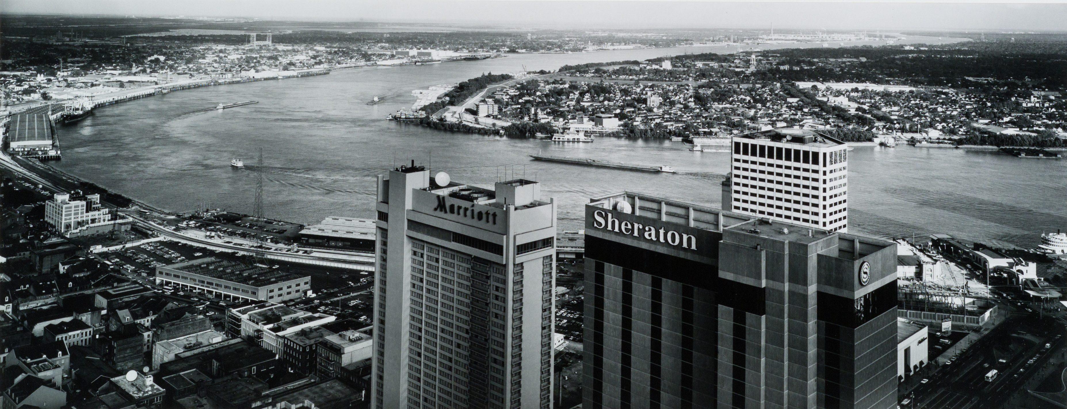 Aerial view of two tall buildings labeled Marriott and Sheraton overlooking a wide river with a cityscape in the background. The winding river separates the buildings from a lush, developed area across the water.