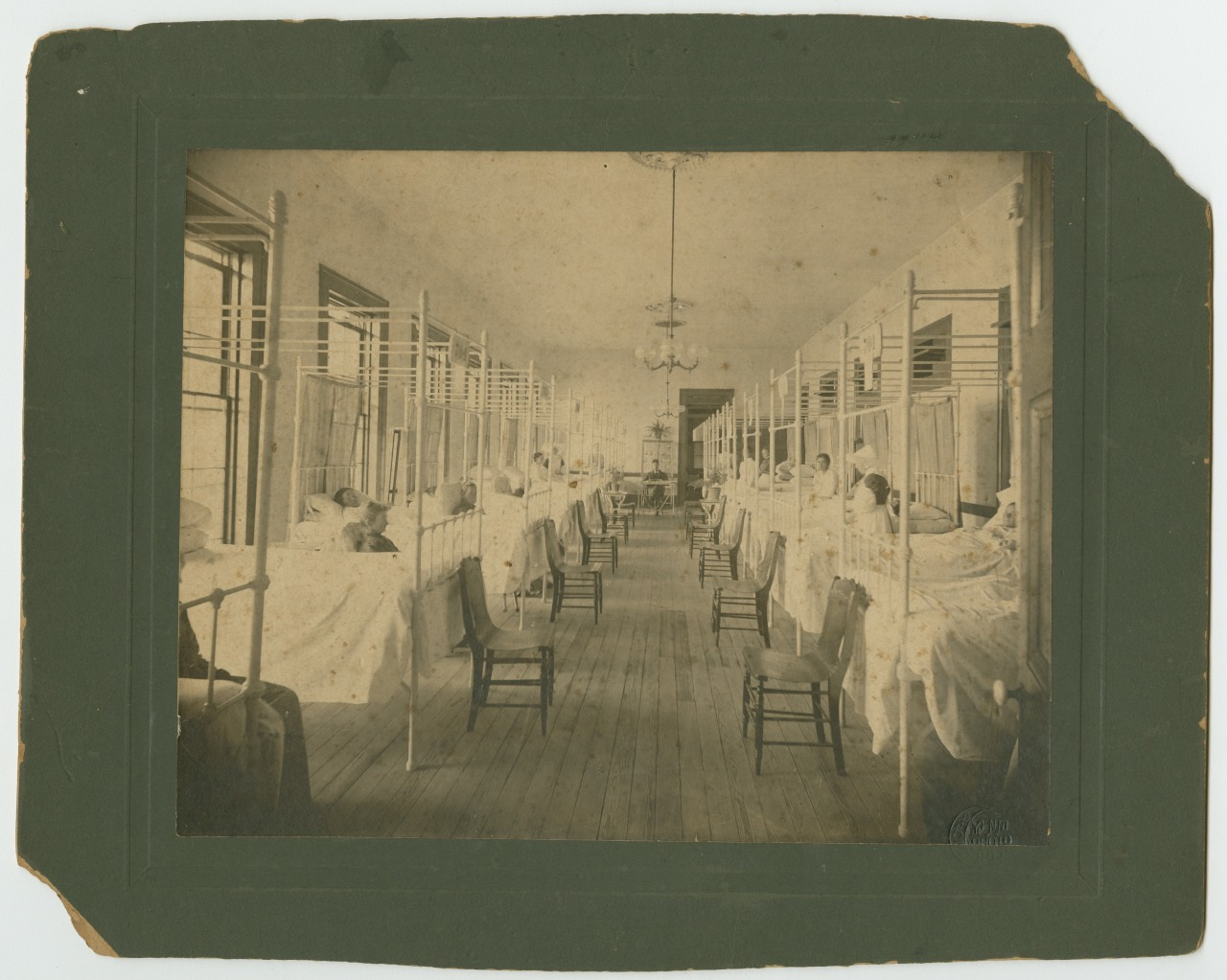 A sepia-toned image of a long, narrow hospital ward with rows of beds on both sides. Patients are lying in the beds, separated by curtains. The room has wooden floors and chairs beside each bed. A chandelier hangs from the ceiling.