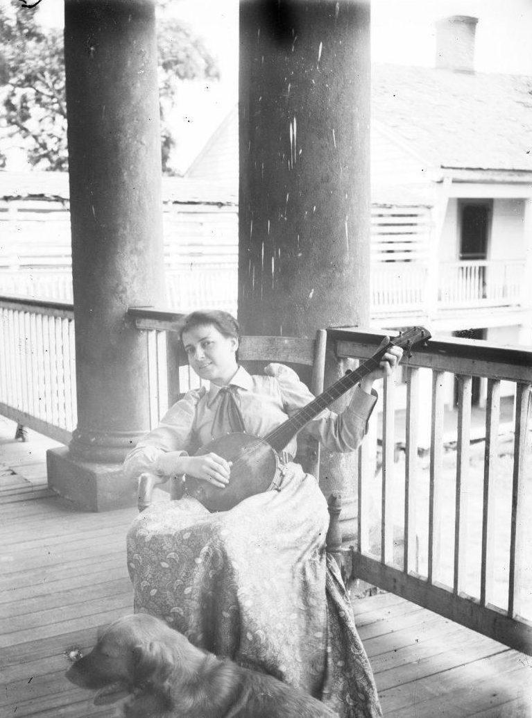 A person sits on a porch playing a banjo. They are dressed in vintage clothing with a long skirt and a ribboned top. A dog lies at their feet. The porch has large columns, and a wooden building is visible in the background.
