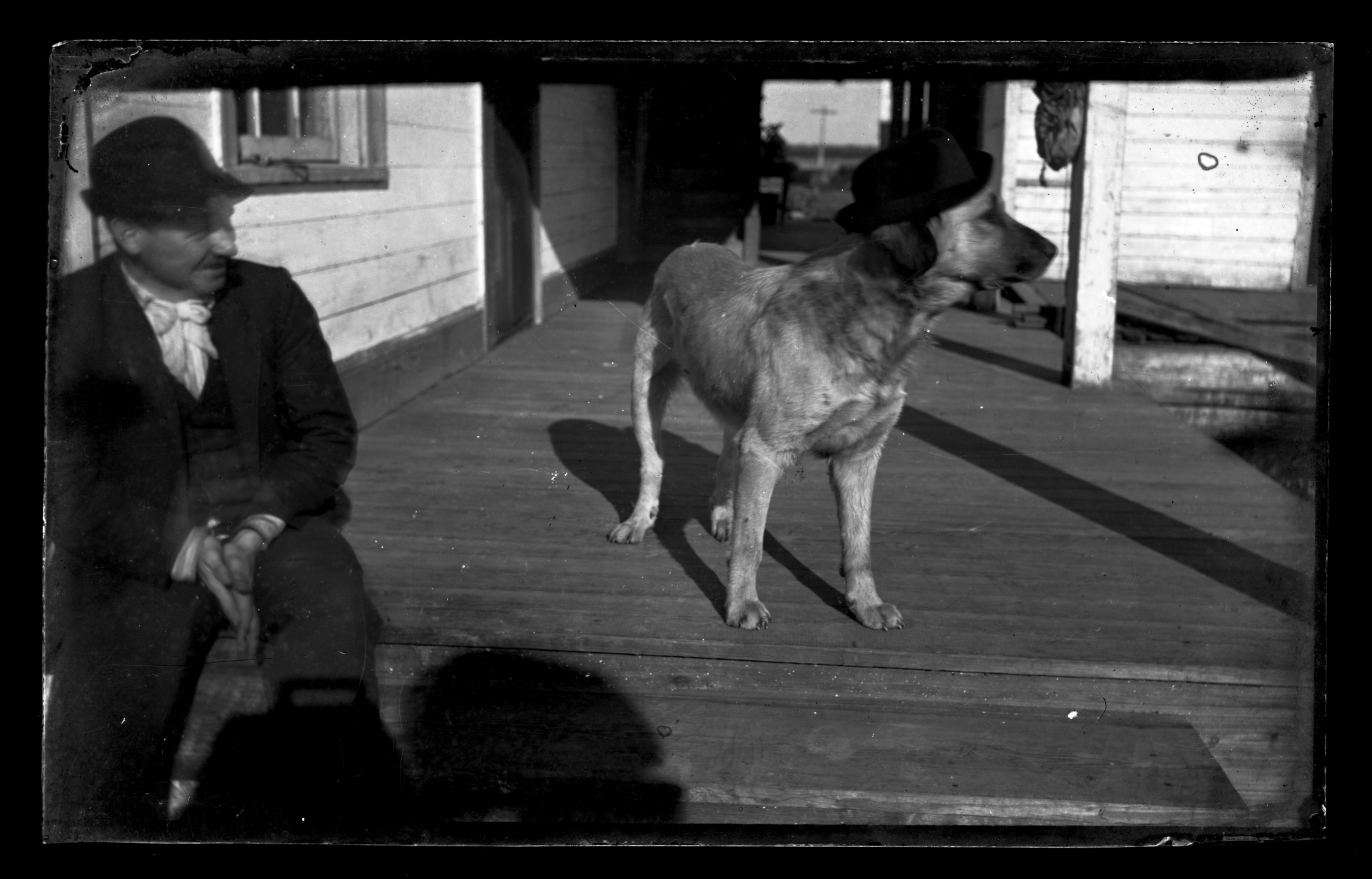 A black and white photo of a man and his dog on a porch. Both are wearing matchin bowler hats.
