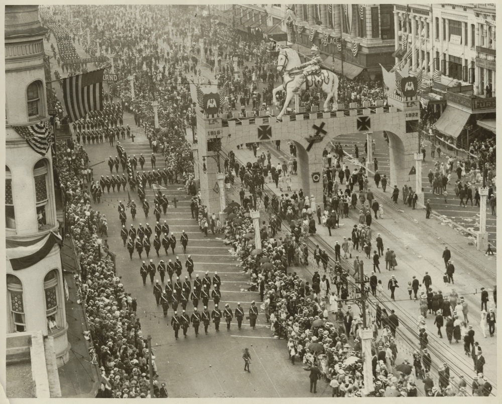 A black and white aerial view of a parade on a wide city street. Marching bands with uniformed members move in disciplined rows. Crowds line the sidewalks, and a decorated arch with horse statues spans the street. Buildings flank both sides.