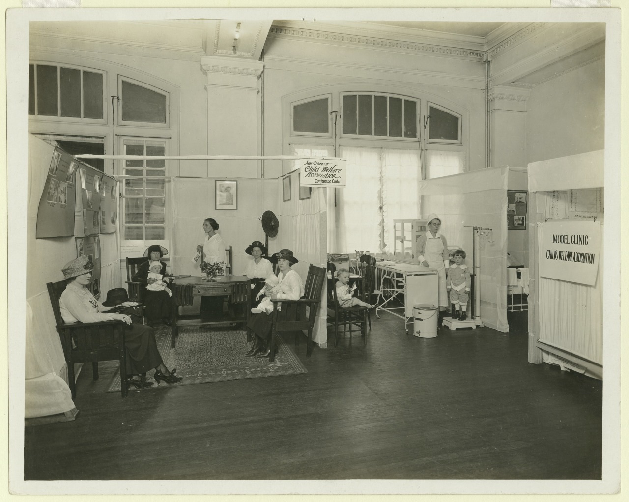 A vintage photo of a clinic with multiple women and children seated or standing. Two nurses are attending to the patients. Signs read Child Welfare Association and Model Clinic. The room has high ceilings and large, curtained windows.