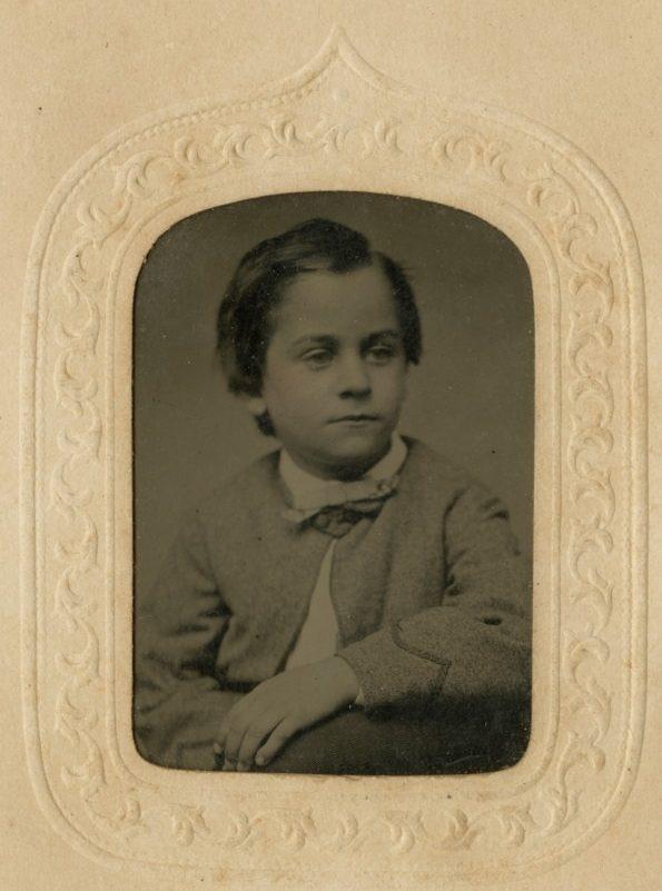 Sepia-toned vintage photograph of a young child with short, dark hair, wearing a suit and bowtie. The image is framed in an ornate, decorative border.