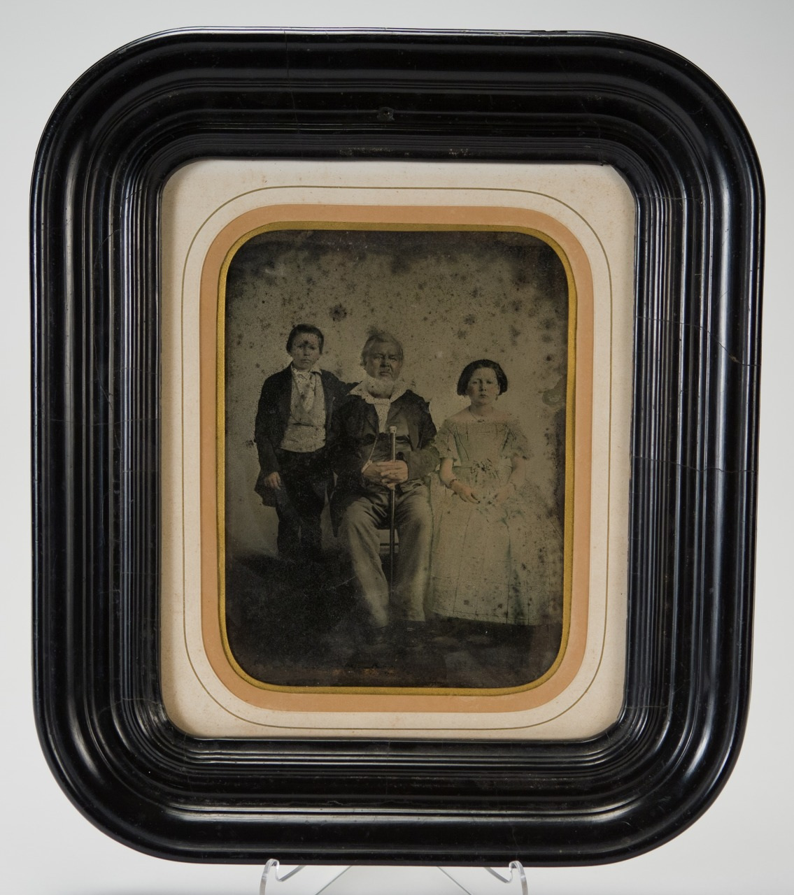 A vintage framed photograph shows three people posing together. The center person is seated with a cane. They are flanked by two others standing; one on each side. The image has a sepia tone and the frame is ornate black with rounded edges.