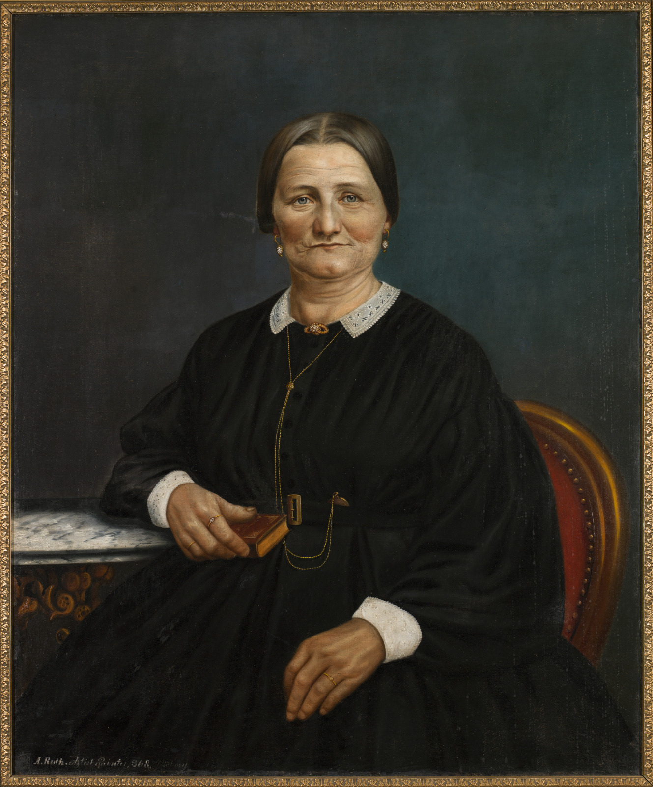 A seated elderly woman wearing a black dress with a white collar holds a small book in one hand. A gold chain hangs from her neck. She sits in a chair beside a table covered with a patterned cloth. The background is dark and plain.