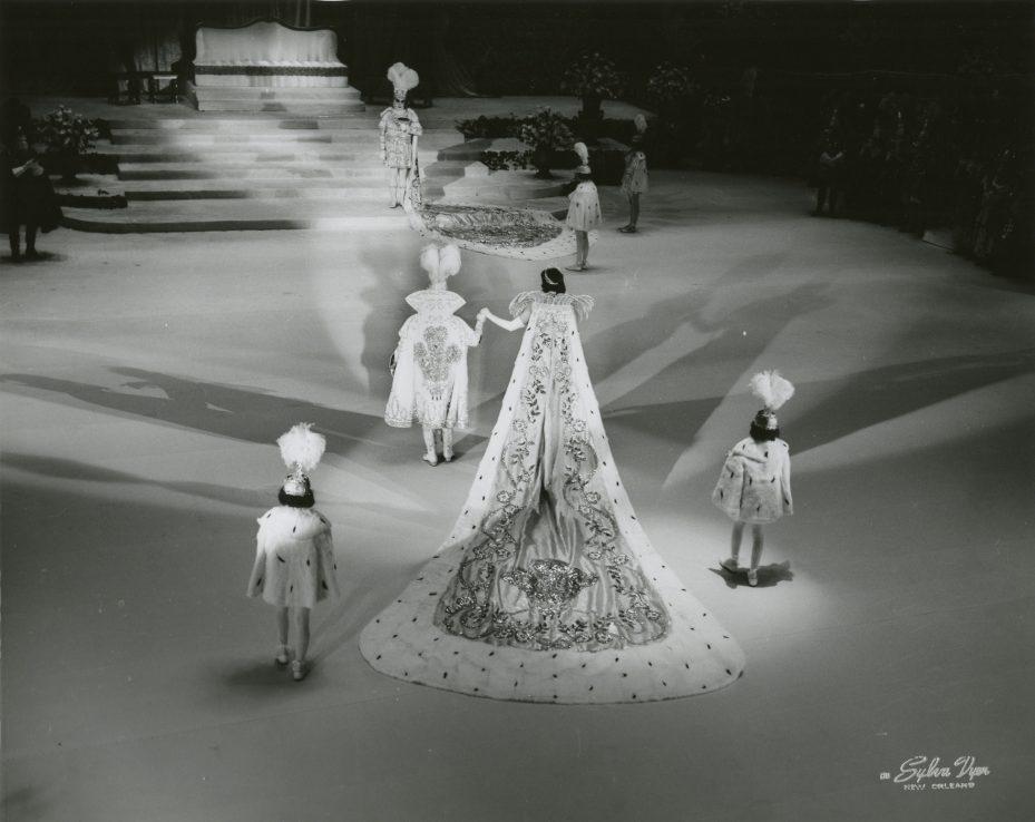 A fashion show features a model in an elaborate gown with a long, embroidered train. The scene is set on a stage with steps in the background. Three people in matching outfits and feathered hats walk alongside.