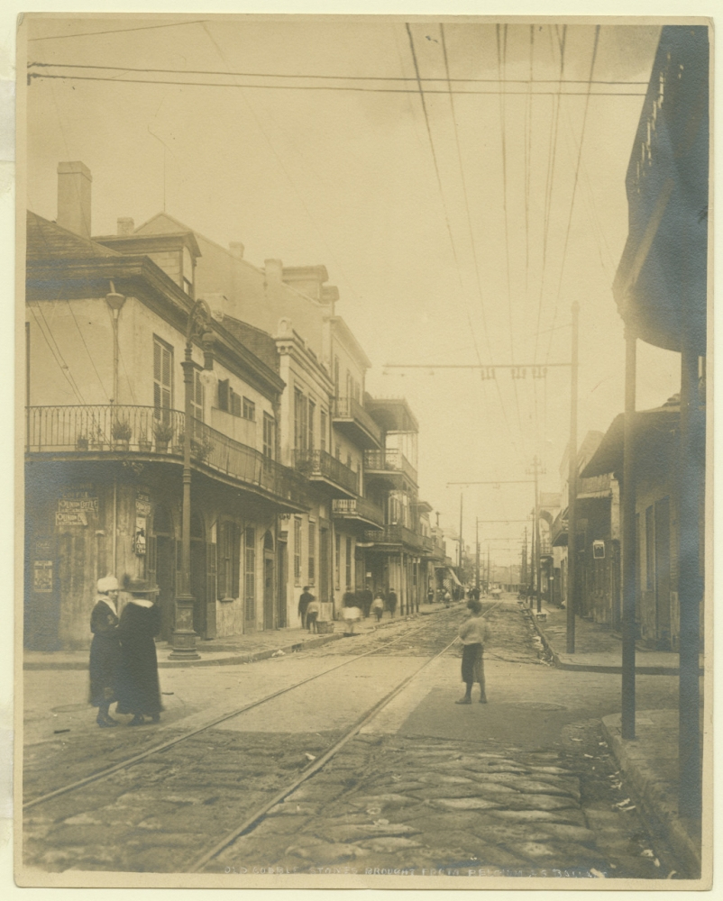 A sepia-toned historical photo of a street with tram tracks, lined with old buildings featuring balconies. People in early 20th-century attire walk along the sidewalk, and a child stands in the road, looking down the street.