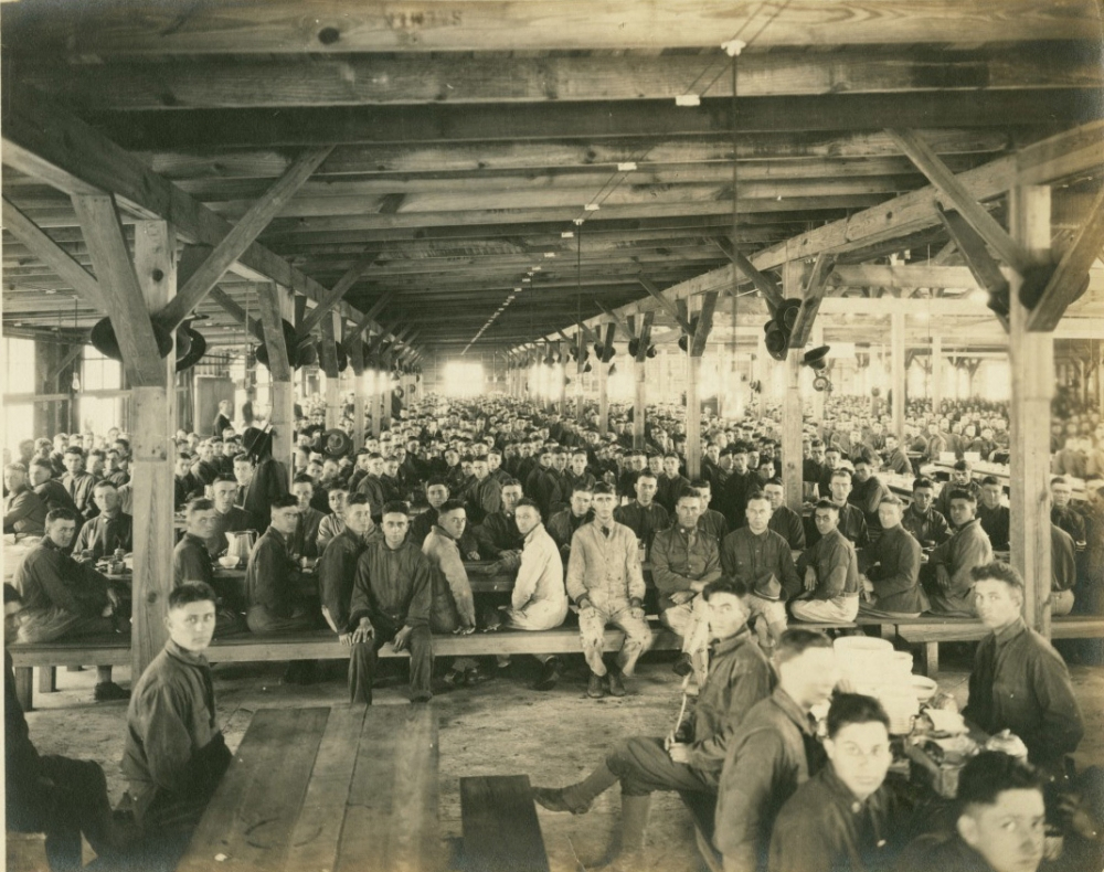 A large group of soldiers sits at long wooden tables in a spacious hall, facing the camera. The space is filled with rows of seated men under a wooden beam ceiling, suggesting a military dining area in historical times.