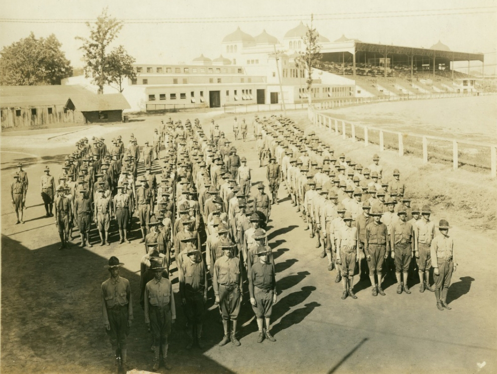 A black and white photo shows a large group of soldiers in uniform standing in formation on an open field. In the background are buildings and a grandstand, with some trees visible. The scene appears to be in a military or training camp.