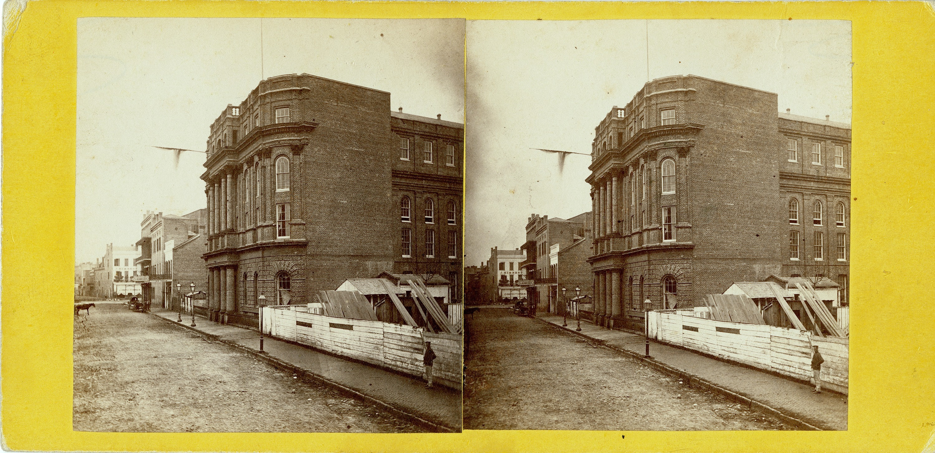 A sepia-toned stereoscopic image of a large brick building on a corner, surrounded by a wooden construction fence. The street is unpaved and a few pedestrians are visible. The background has more buildings and a cloudy sky.