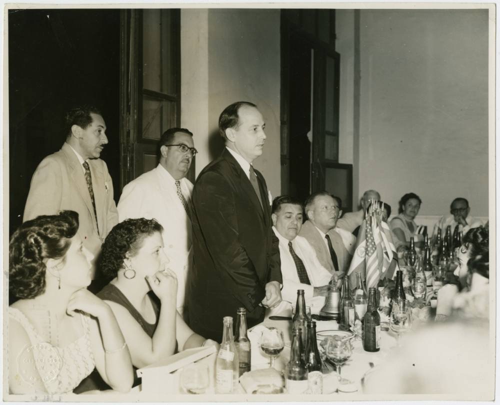 A black-and-white photo of people gathered around a long dining table with bottles and glasses. A man in a suit stands while others sit, listening. The setting appears formal with both men and women present.