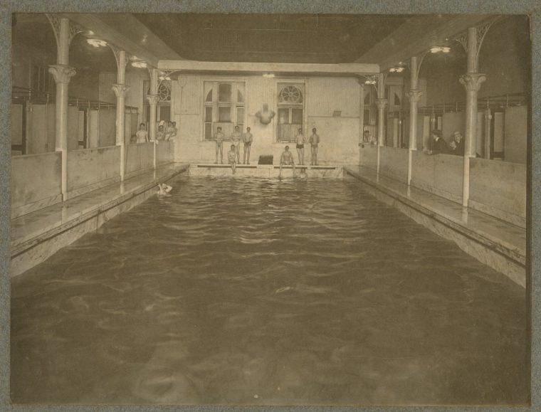 A vintage indoor swimming pool with several people standing at the pools edge. The room has ornate columns and windows, and the pool water appears dark. The atmosphere is historical and the image is in sepia tones.