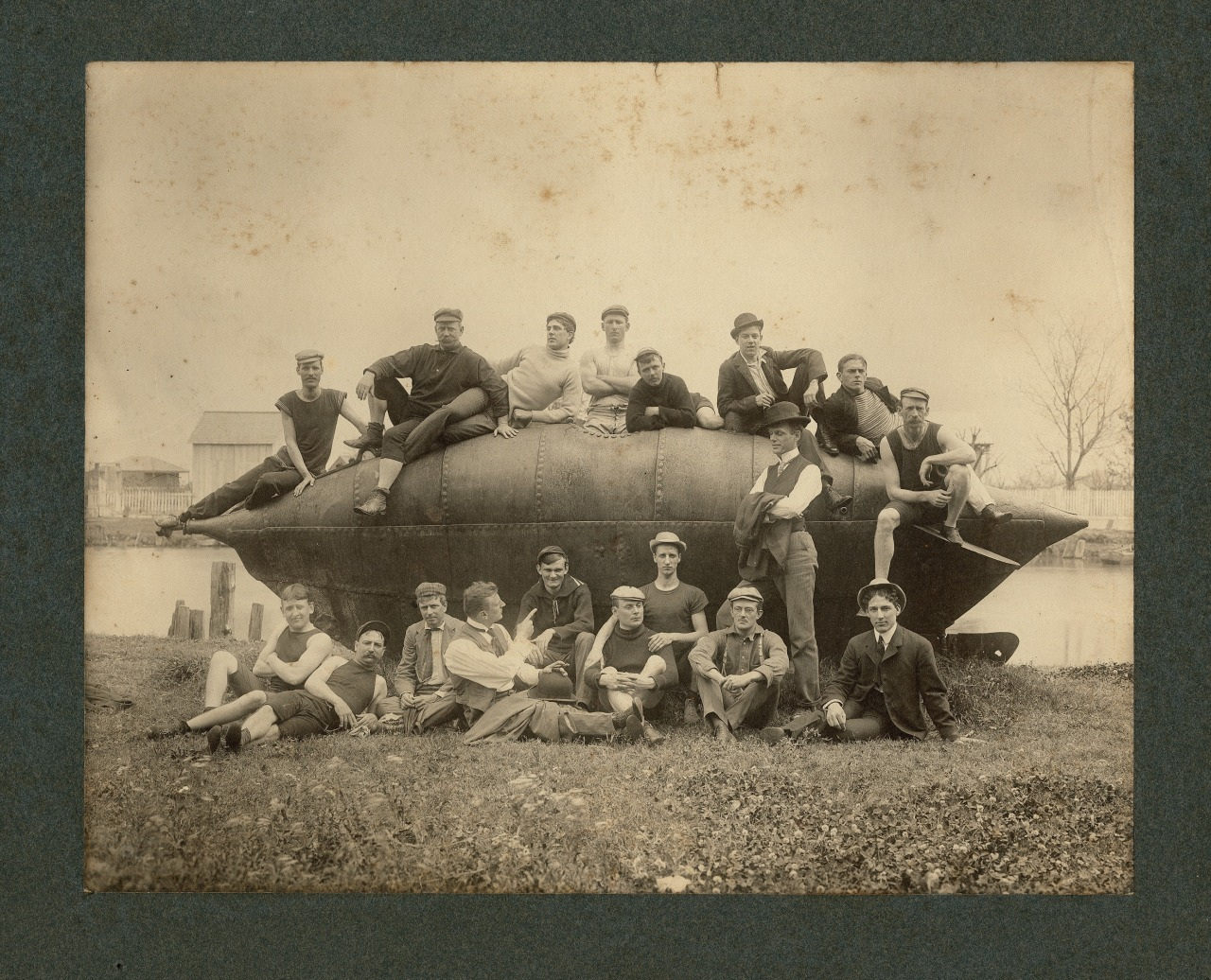 A vintage sepia-toned photo shows a group of men wearing casual, late 19th to early 20th-century clothing and sailor caps. They are gathered and seated on a large metal structure near a waterfront, with trees and buildings in the background.