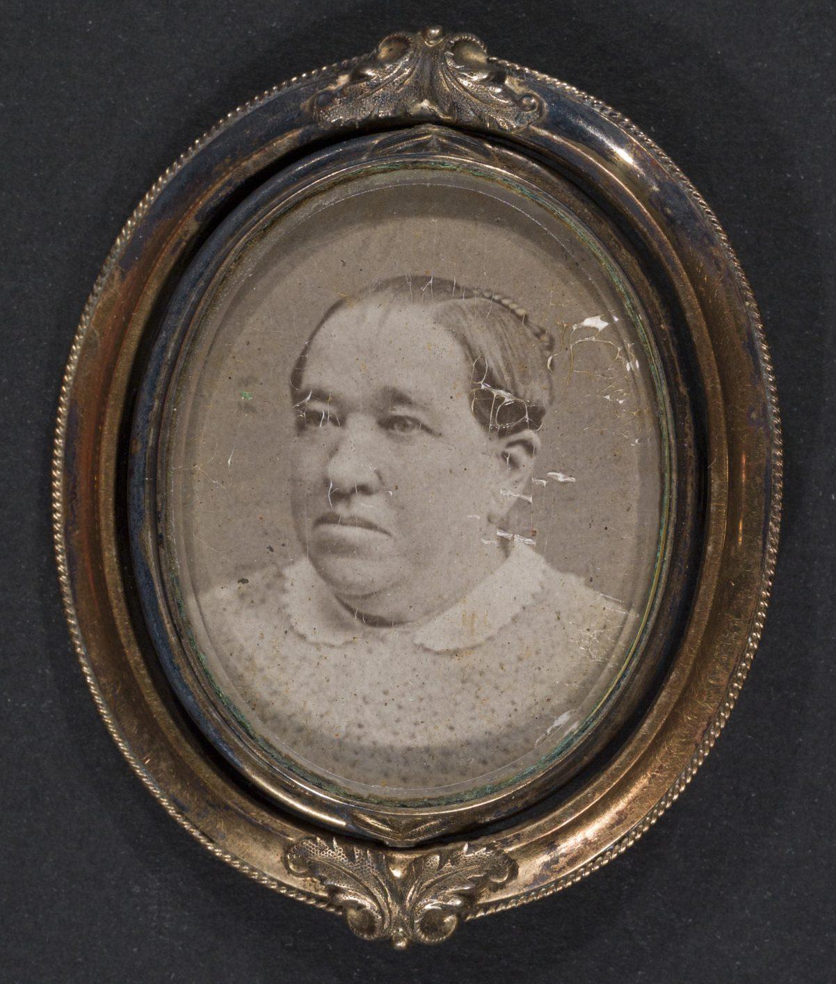 An old sepia-toned photograph in an oval ornate frame shows a middle-aged person with a serious expression, wearing a light-colored garment with a lace collar. The photo has some wear and scratches.