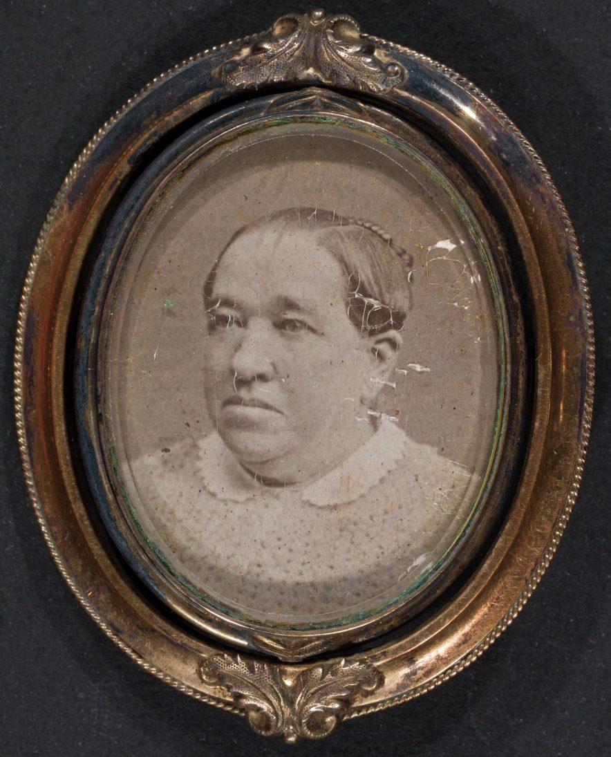 A vintage oval portrait photograph of a person with a neutral expression, encased in an ornate gold frame. The person is wearing a light-textured garment. The photo shows some signs of aging and damage.