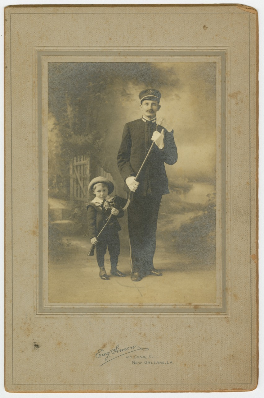 A sepia-toned vintage photograph shows a man in a military uniform and a young boy standing beside him. Both are holding canes, wearing hats, and facing the camera. A garden gate and foliage are visible in the blurred background.
