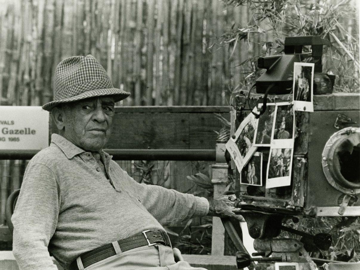 An elderly man wearing a hat sits next to a vintage camera adorned with multiple photographs. A sign with partial text is visible in the background. The scene is outdoors, with plants around. The image is in black and white.