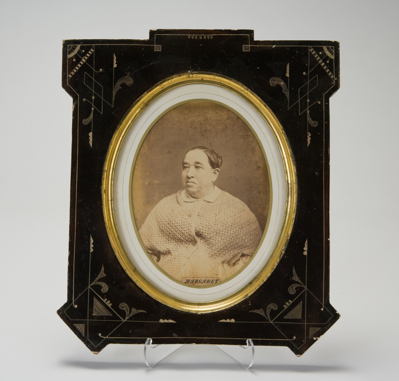 A vintage framed sepia-toned photograph of a person wearing a light-colored shawl. The ornate frame is black with decorative patterns and a gold inner border. The photograph is displayed on a clear stand.