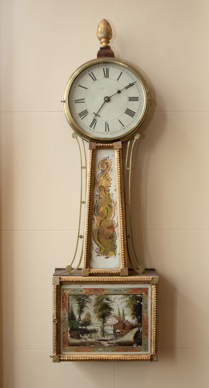 A vintage pendulum wall clock with a round face, Roman numerals, and decorative gold accents. The lower section features a painted scene of a house by a river surrounded by trees.