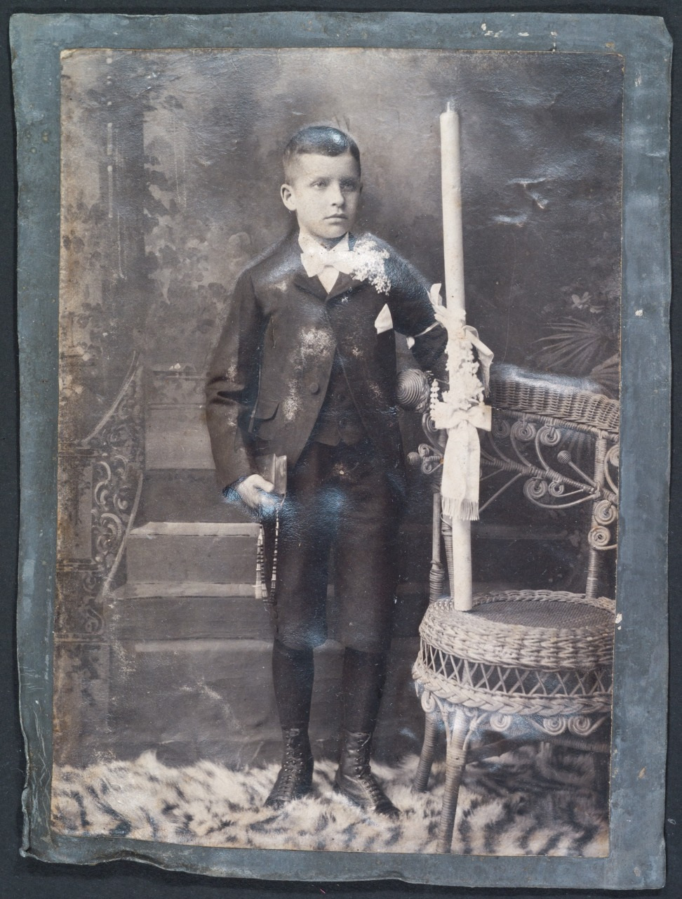 Vintage photo of a young boy in formal attire, standing next to a wicker chair and holding a candle. He is posed on a patterned rug with a decorative backdrop featuring steps and foliage.