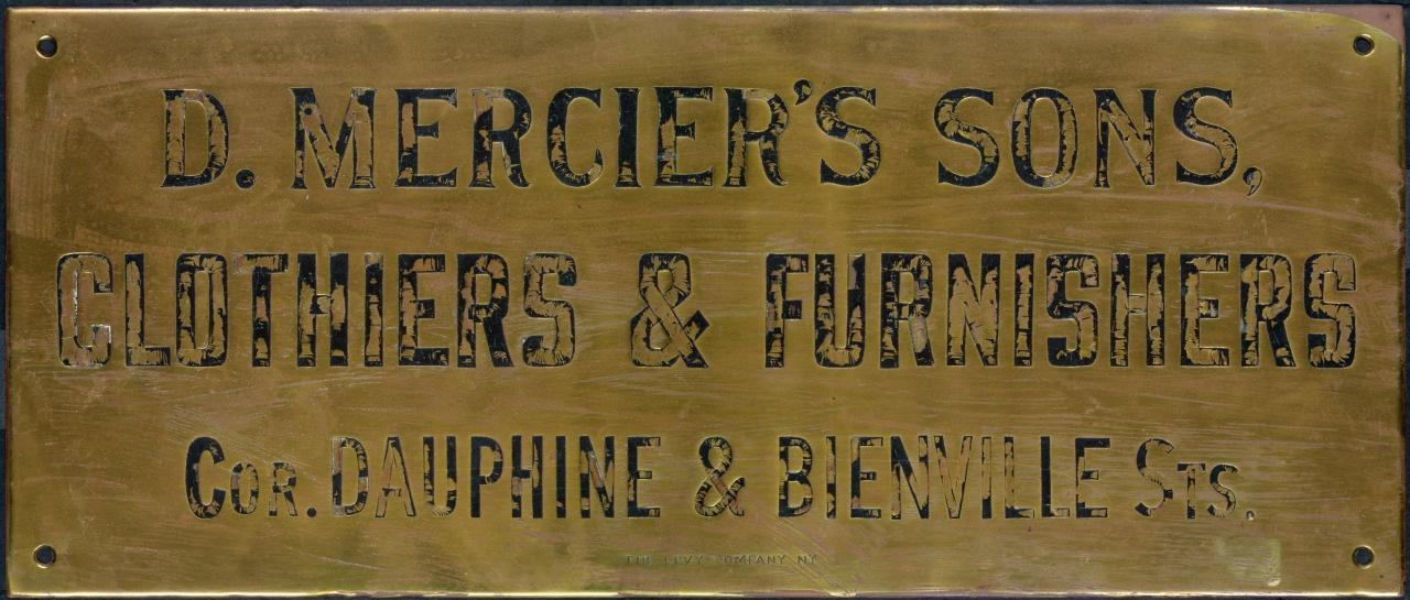 A vintage brass sign reads D. Merciers Sons, Clothiers & Furnishers, Cor. Dauphine & Bienville Sts. The text is engraved in a bold, classic font, with some patina and wear evident on the surface of the sign.