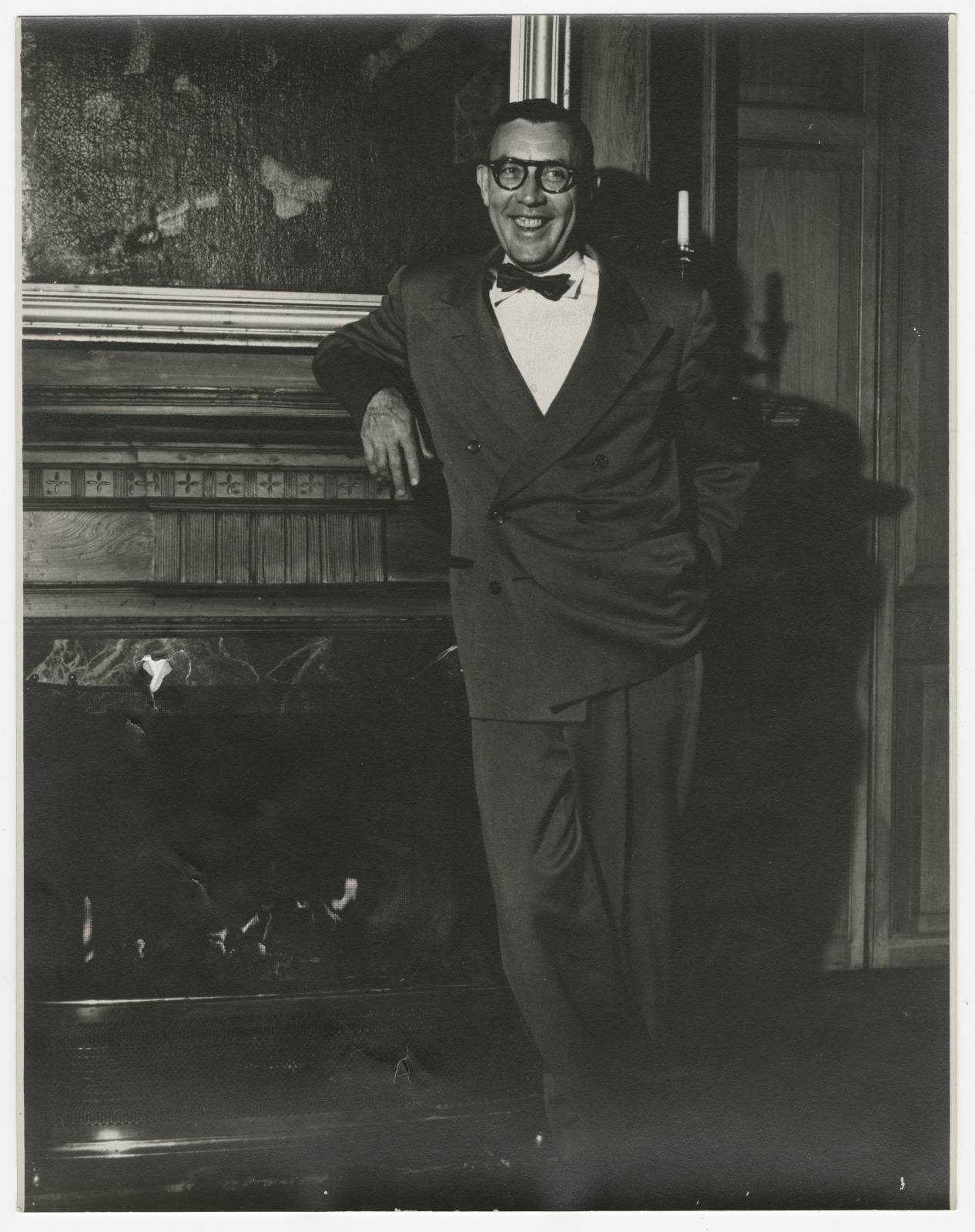 A man in a dark suit and bow tie (Boyd Cruise) standing in front of a fireplace.