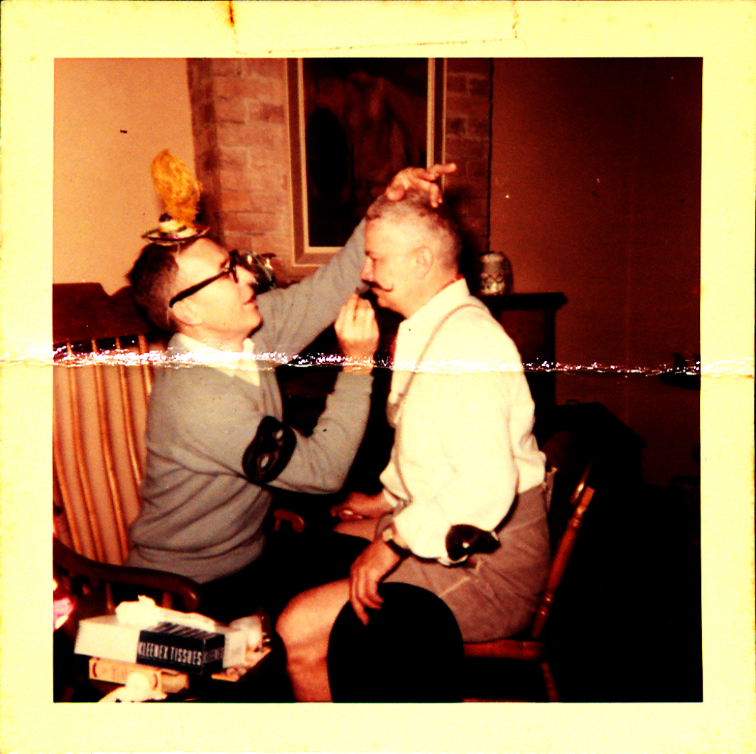 An early color photo of a man in a grey sweater (Boyd Cruise) drawing a mustache on a balding man in a white shirt (Harold Schilke). A brick fireplace is visible in the background.