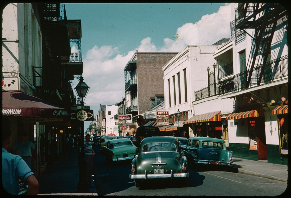 Street scene from a mid-20th-century city, featuring vintage cars, including a green sedan, and storefronts with colorful awnings. Urban architecture with fire escapes is visible under a partly cloudy sky. Pedestrians walk along the sidewalk.